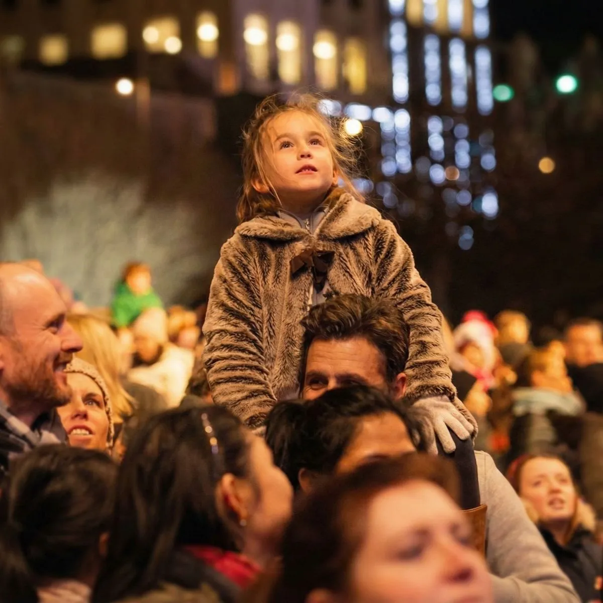 A young child wearing a colorful wool hat looking up in awe at sparkling lights during the Bairns Afore Edinburgh Hogmanay event.