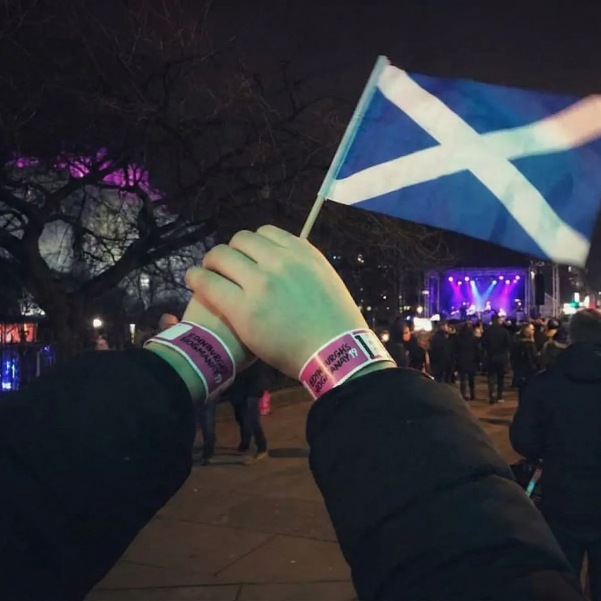 A close-up of a colorful Hogmanay event wristband on a wrist with blurry Christmas market lights in the background.