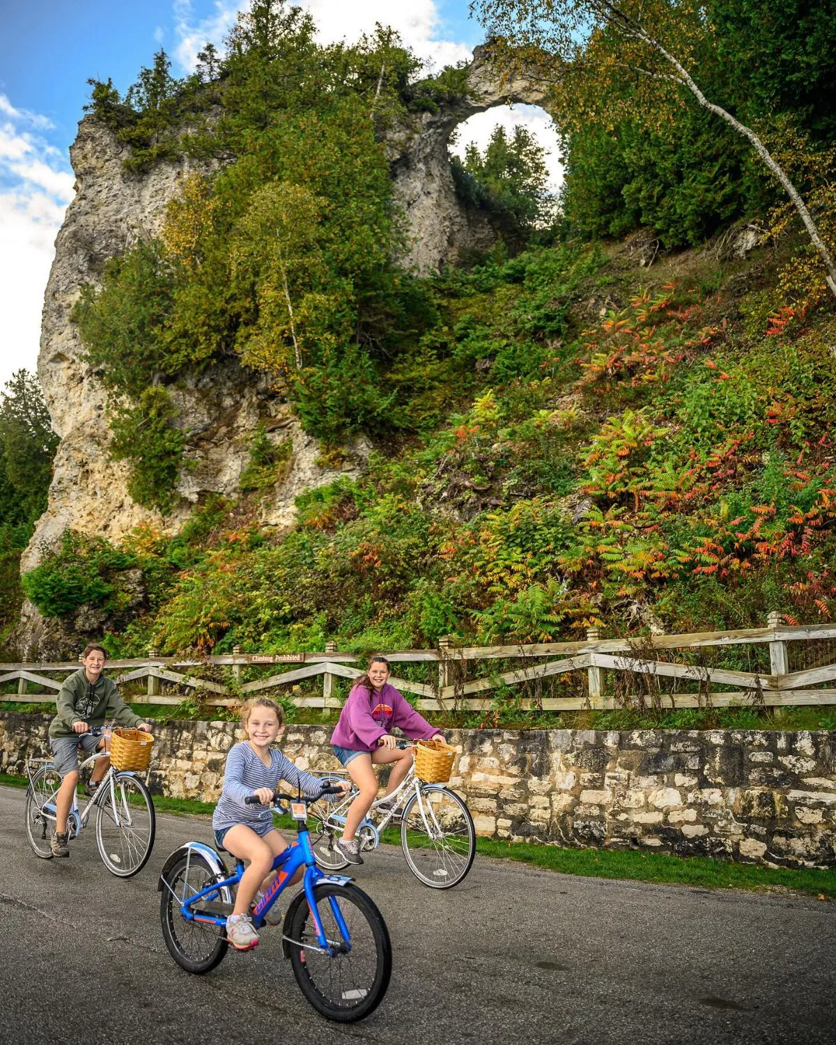 A group of people riding bicycles on a paved coastal path next to turquoise water and white rocks on Mackinac Island.