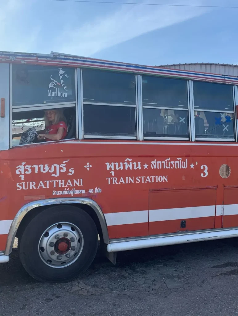 A classic open-air songthaew taxi drives along a scenic coastal road surrounded by lush tropical greenery on a sunny day in Koh Phangan, Thailand.
